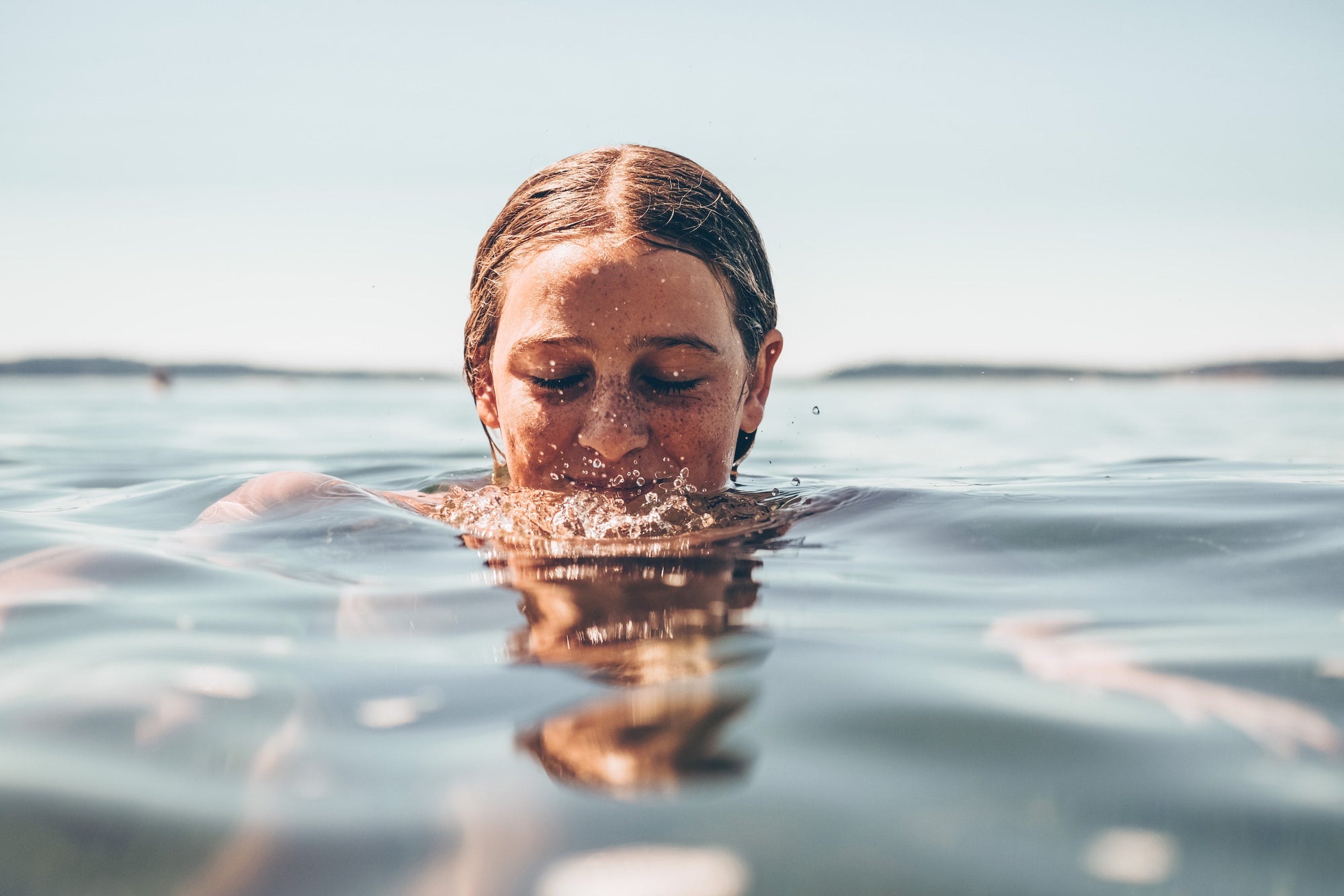 A person with wet hair and freckles partially submerged in calm water with eyes closed, blowing bubbles on the water surface.