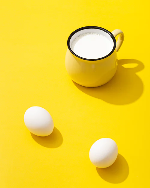 Yellow mug filled with milk and two white eggs casting shadows on a bright yellow surface.