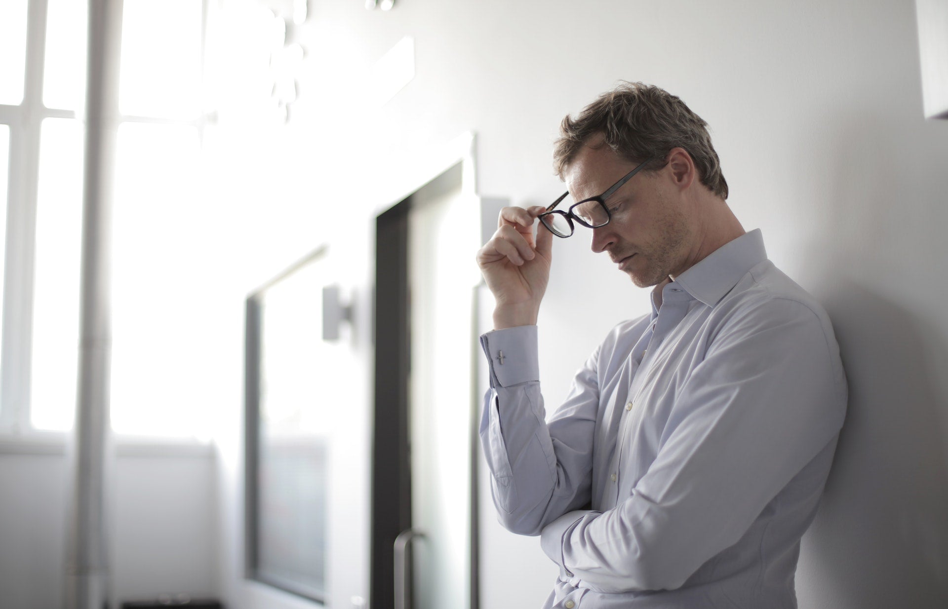 Man in light blue shirt holding glasses and leaning against a wall, appearing stressed or contemplative in a bright room