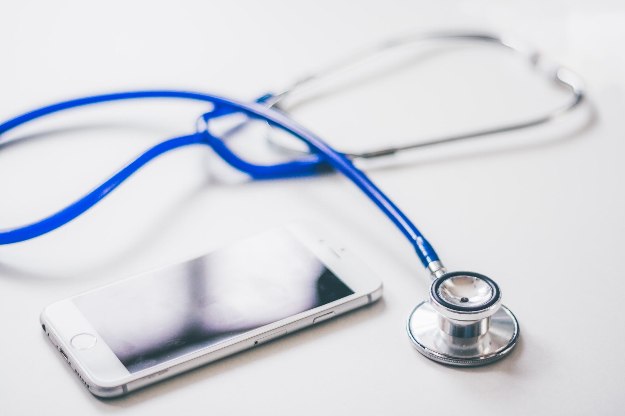 Blue stethoscope lying next to a white smartphone on a white surface