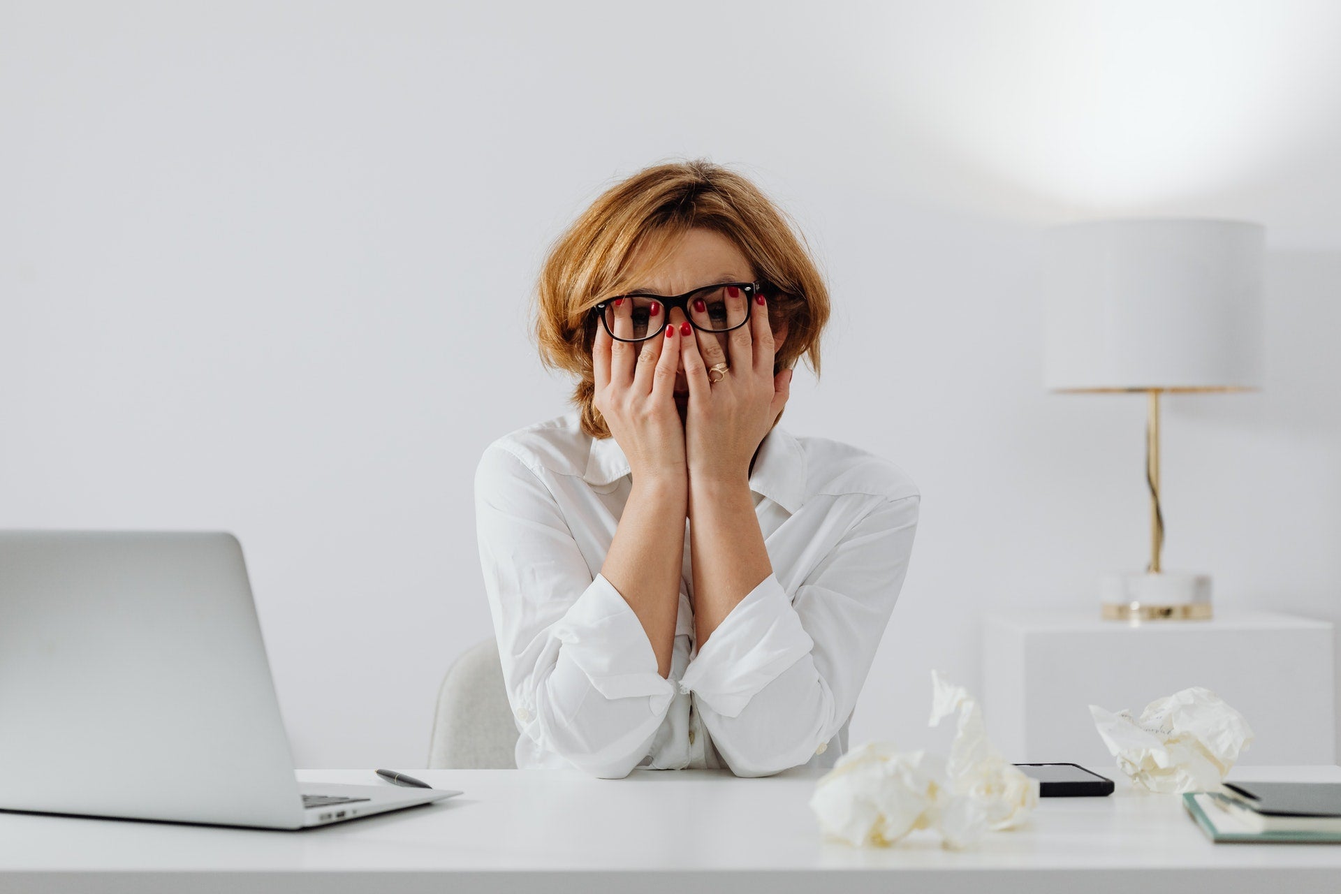 Stressed woman with glasses covering her face at a desk with crumpled papers, laptop, and lamp, illustrating anxiety and serotonin-related stress.