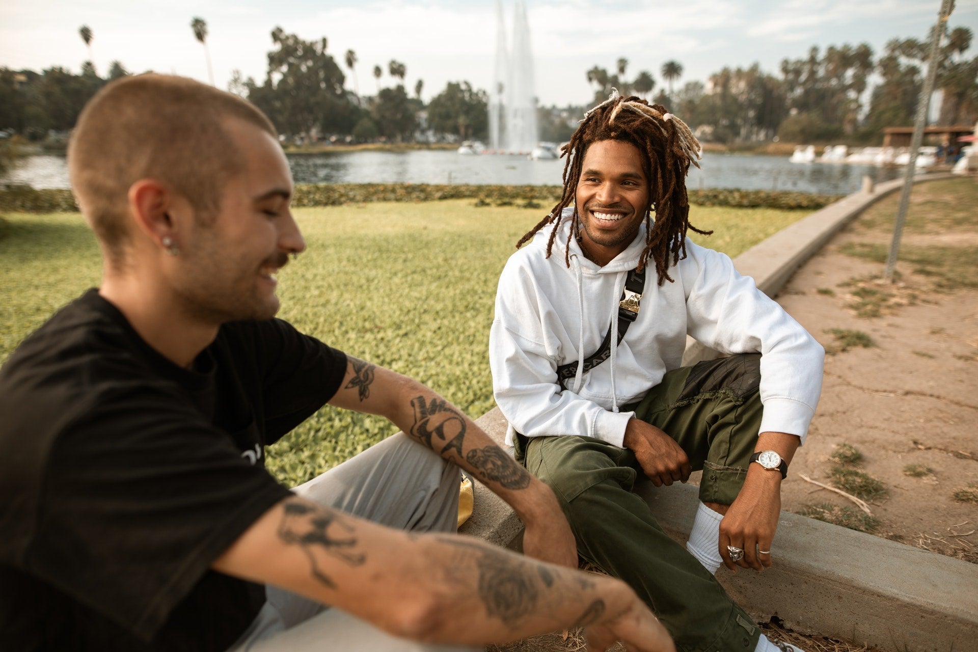 Two men sitting and smiling outdoors near a lake with a fountain and palm trees in the background, one wearing a white hoodie and the other a black t-shirt with tattoos on his arms.