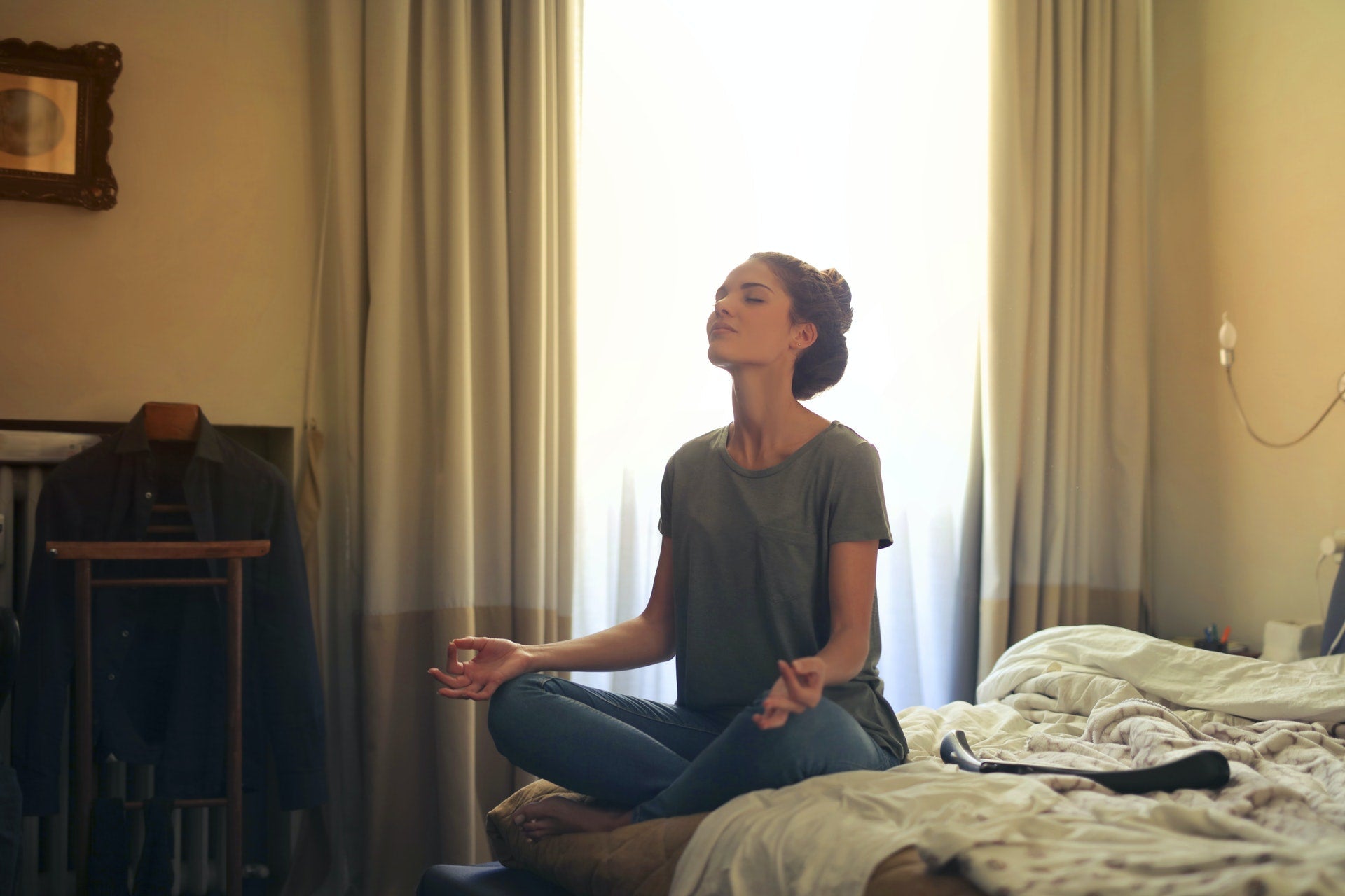Woman sitting cross-legged on a bed meditating with eyes closed in a softly lit bedroom with curtains and clothes hanging in the background