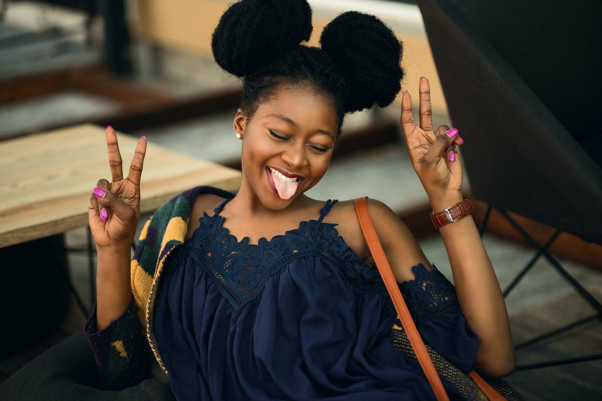 Young woman with two afro puff hairstyles smiling with eyes closed and sticking out her tongue, making peace signs with both hands, wearing a navy blue lace top and a brown shoulder bag.