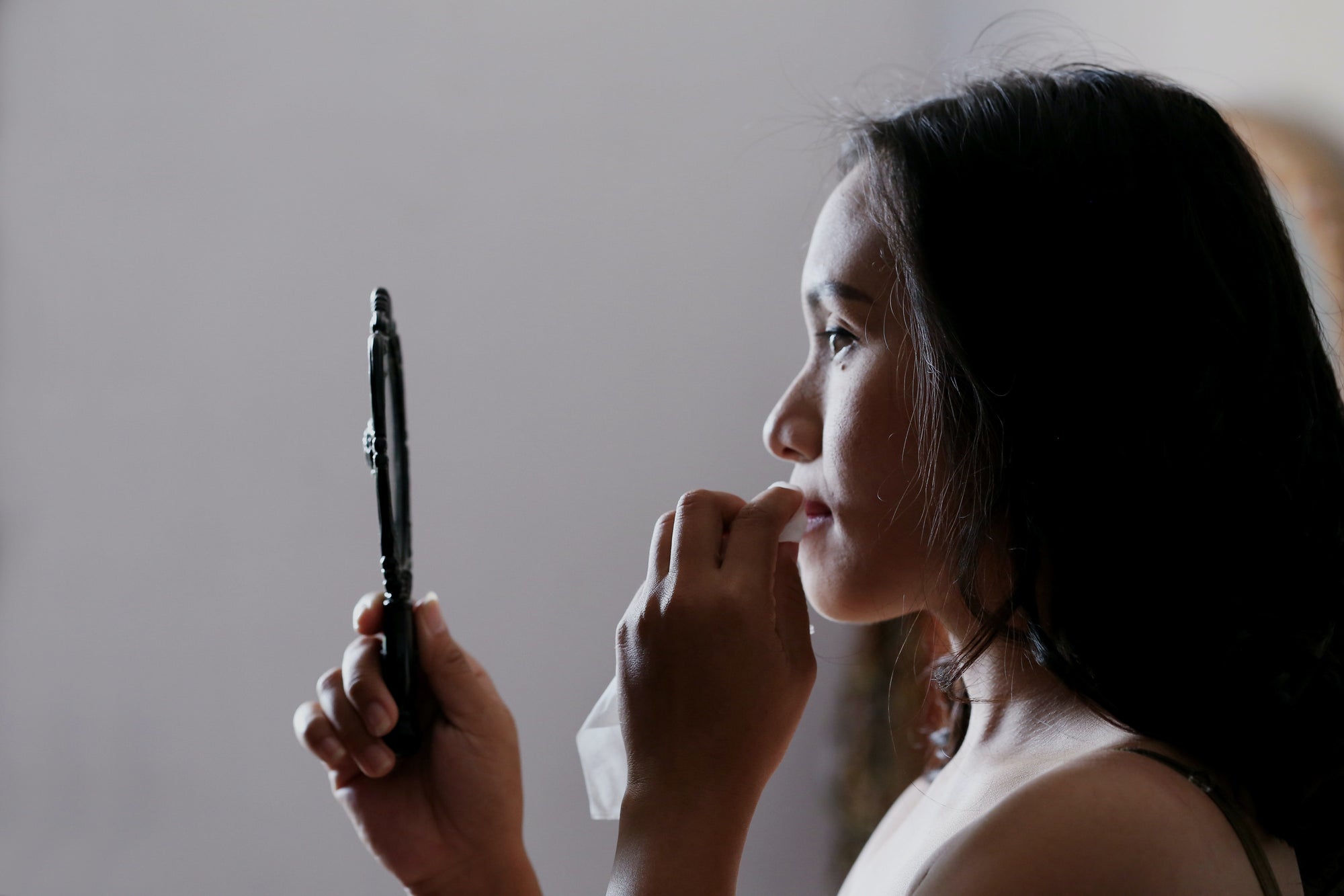 Woman looking into a handheld mirror while holding a tissue near her mouth, in profile view
