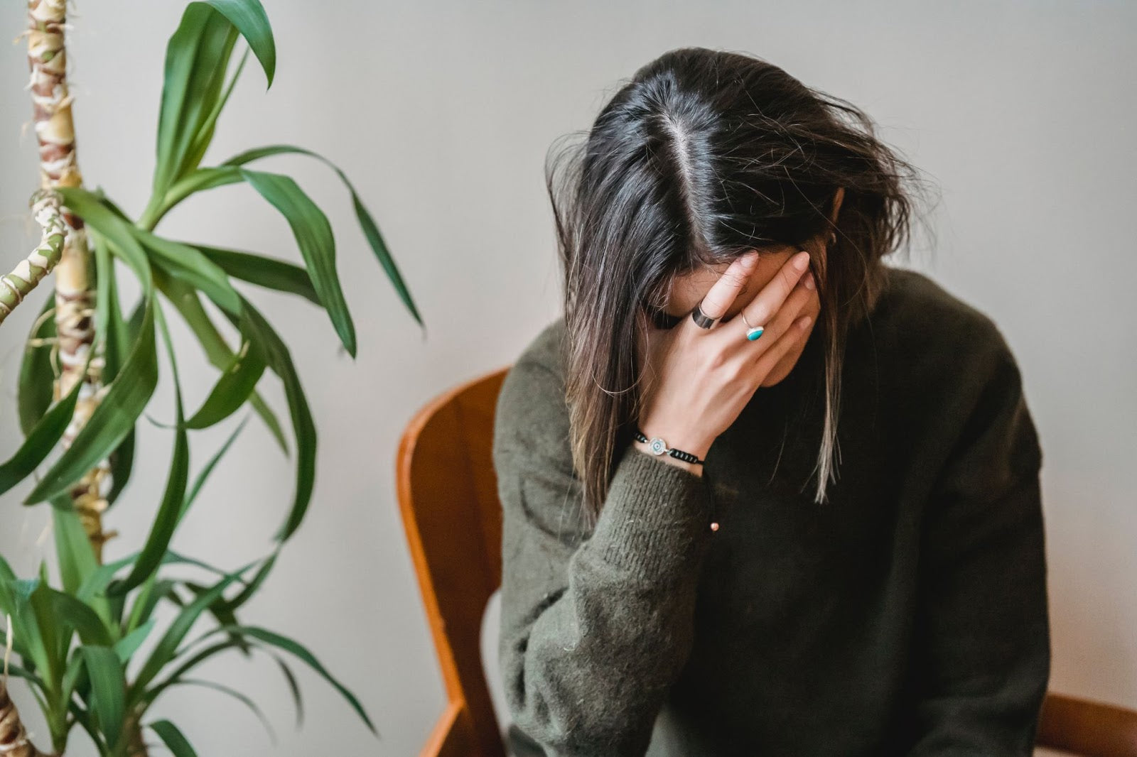 Woman sitting on a chair with her hand covering her face, appearing distressed, next to a green plant.