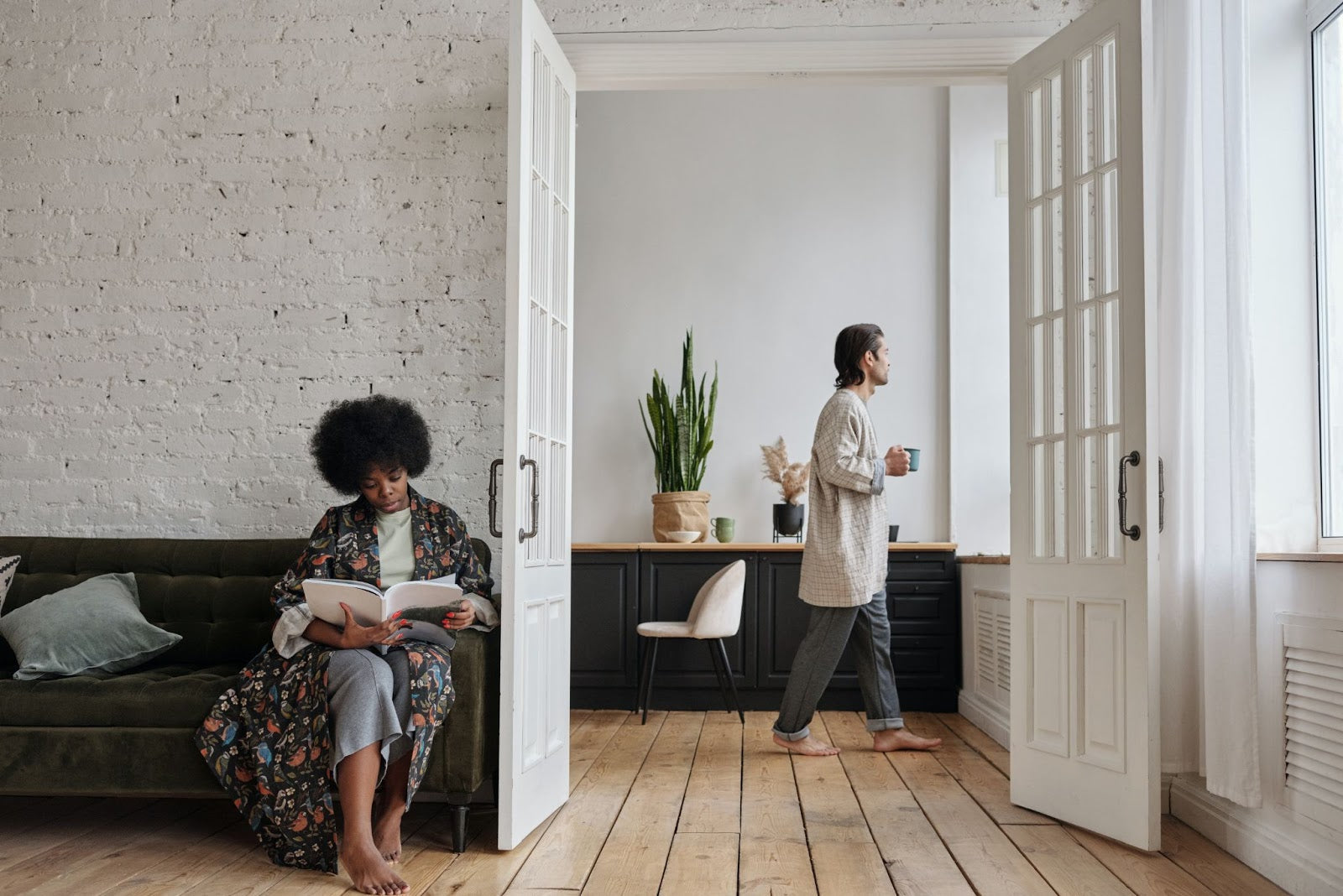 A woman reading a book on a green couch and a barefoot man walking with a cup in a bright room with wooden floors and white walls.