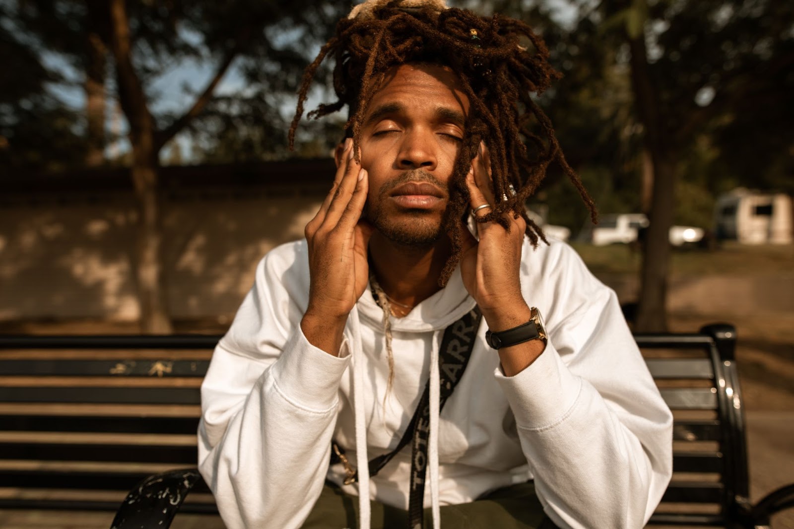 Man with dreadlocks sitting on a bench outdoors, eyes closed and hands on his face, appearing to calm himself.
