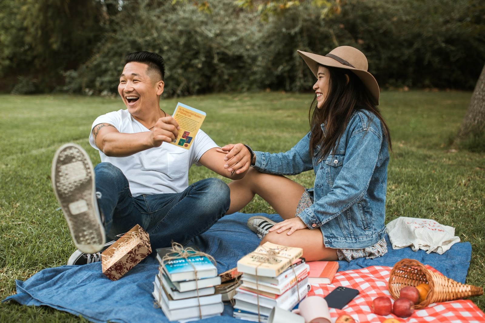 Two people sitting on a picnic blanket outdoors, smiling and holding hands, surrounded by stacks of books and a basket of fruit.