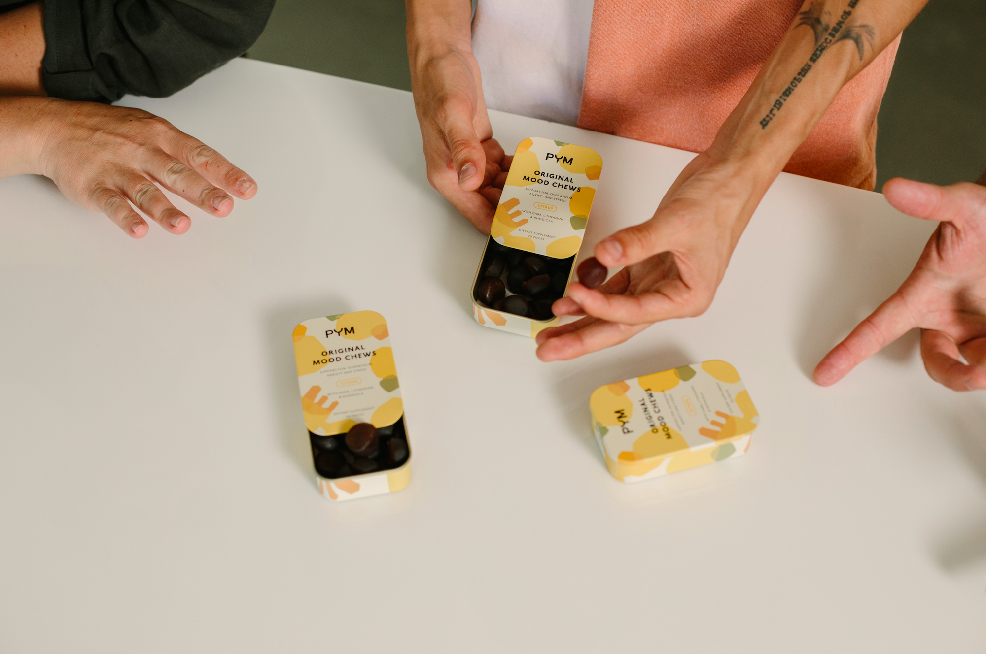 Hands holding and reaching for tins of PYM Original Mood Chews labeled "Original Mood Chews" and "Citrus" on a white table.