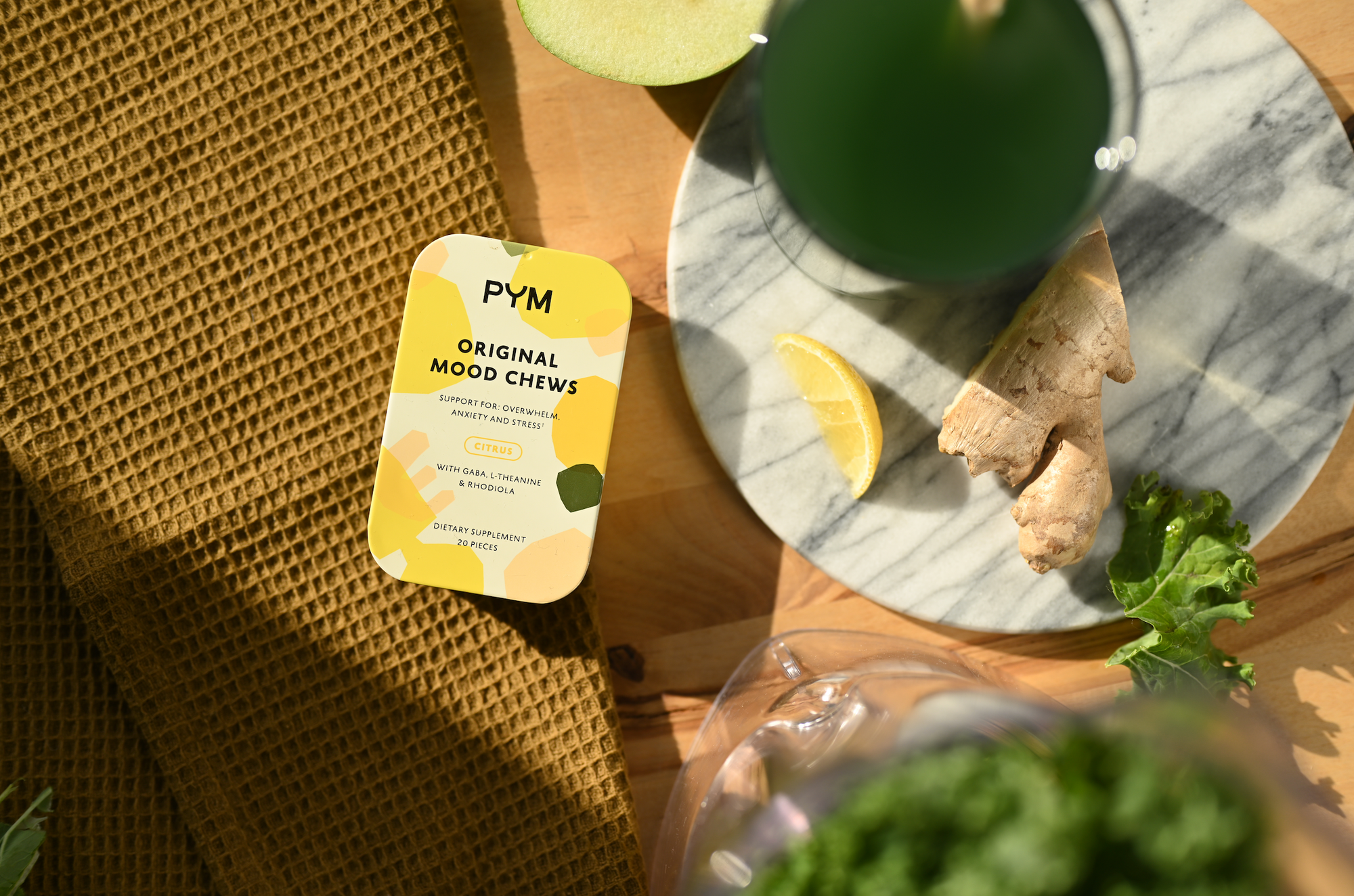 Yellow tin of PYM Original Mood Chews next to a lemon slice, ginger root, and a green drink on a wooden surface.