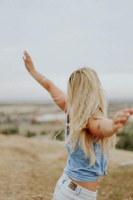 Blonde woman outdoors with arms outstretched, wearing a blue tank top and white shorts, with a blurred natural landscape background.