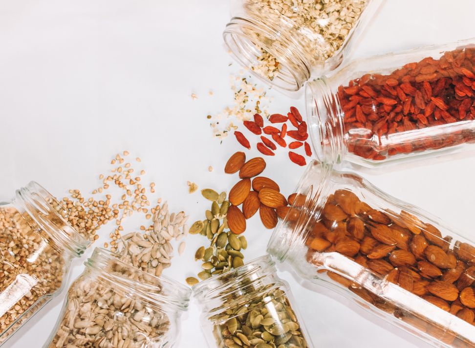 Various nuts and seeds including almonds, pumpkin seeds, sunflower seeds, goji berries, and buckwheat spilled from glass jars on a white background.
