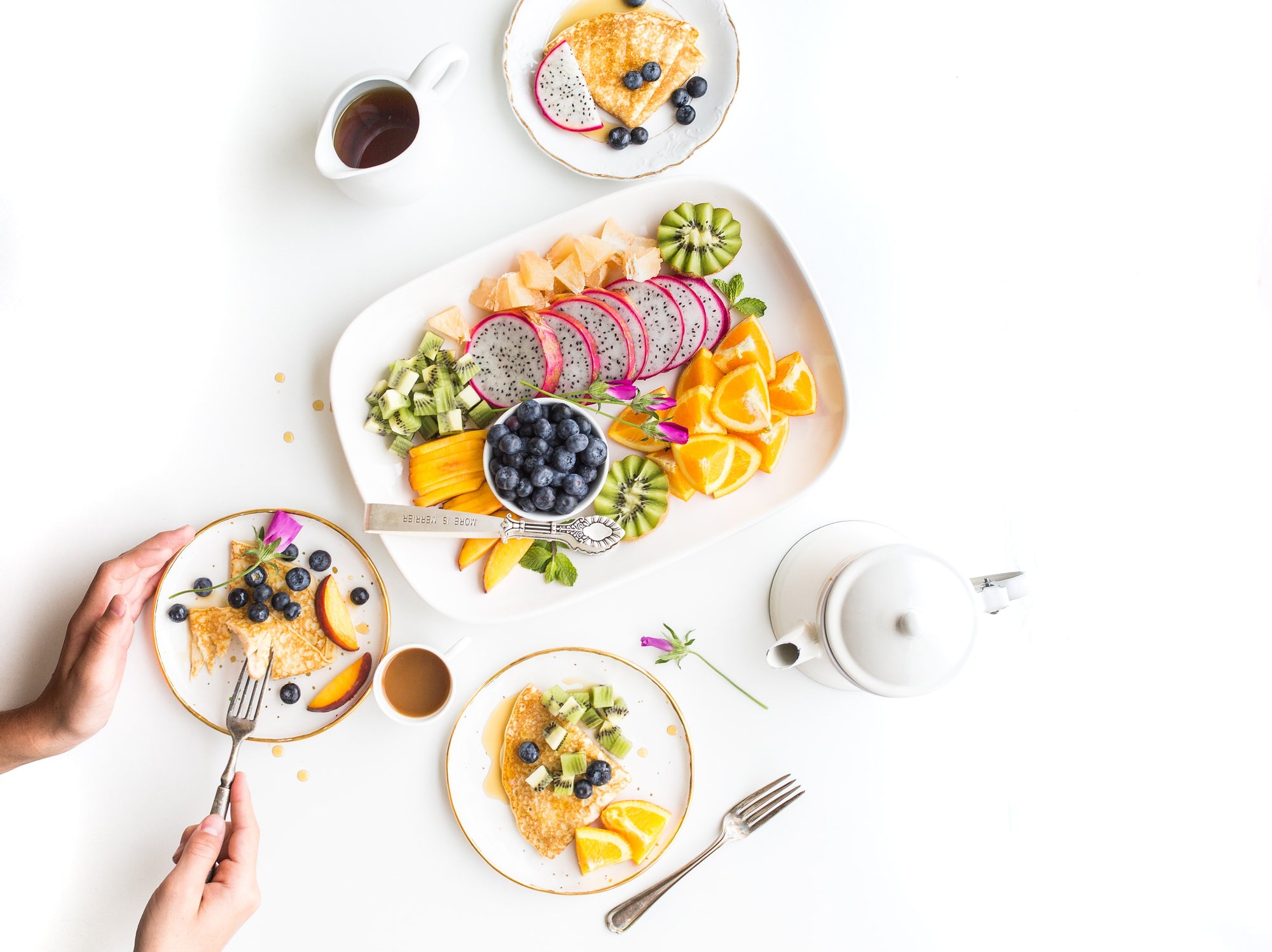 Breakfast spread with pancakes topped with blueberries and kiwi, a platter of sliced dragon fruit, orange wedges, kiwi, and blueberries, accompanied by a teapot, coffee, and tea cups on a white table.