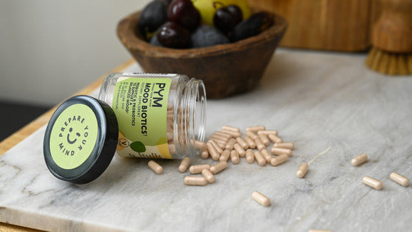Jar of PYMBIOTICS Mood Biotics capsules spilled on a marble surface with a bowl of olives in the background. The jar lid reads 'PREPARE YOUR MIND'.