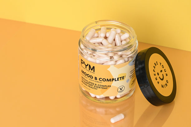 Open jar of PYM Mood B Complete+ supplement capsules with label reading 'PYM Mood B Complete+ Supplement Methylated B-Complex to support everyday good health' and lid text 'Prepare Your Mind'. One capsule lies outside the jar on a reflective surface.