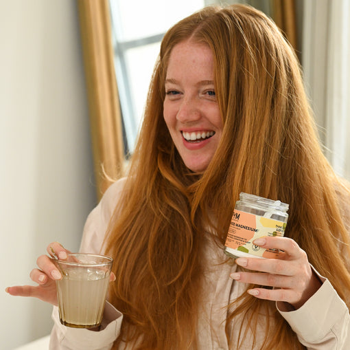 Smiling woman holding a jar of PYM Mood Magnesium sleep supplement and a glass with liquid indoors near a window.