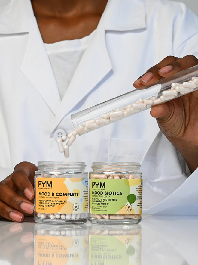 Two jars of PYM dietary supplements labeled 'Mood B Complete' and 'Mood Biotics' with capsules being poured from a test tube by a person in a white coat.