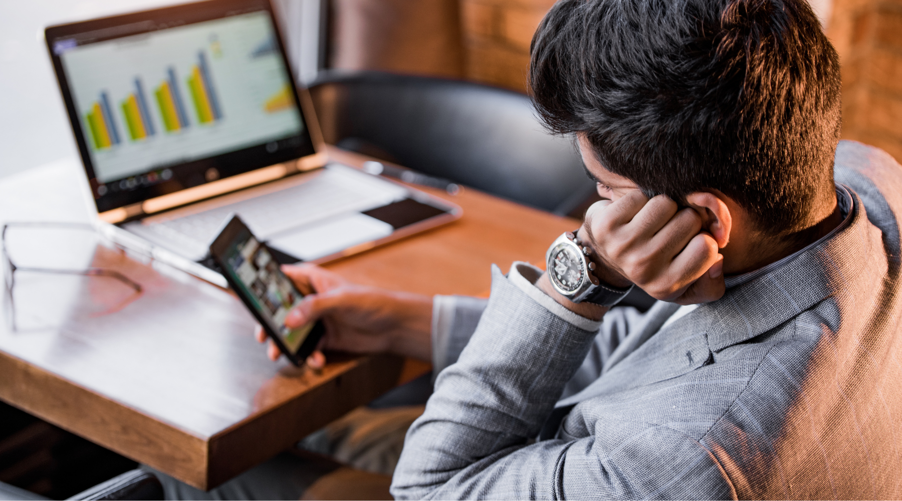 Man in gray jacket looking at smartphone with laptop showing bar chart in background