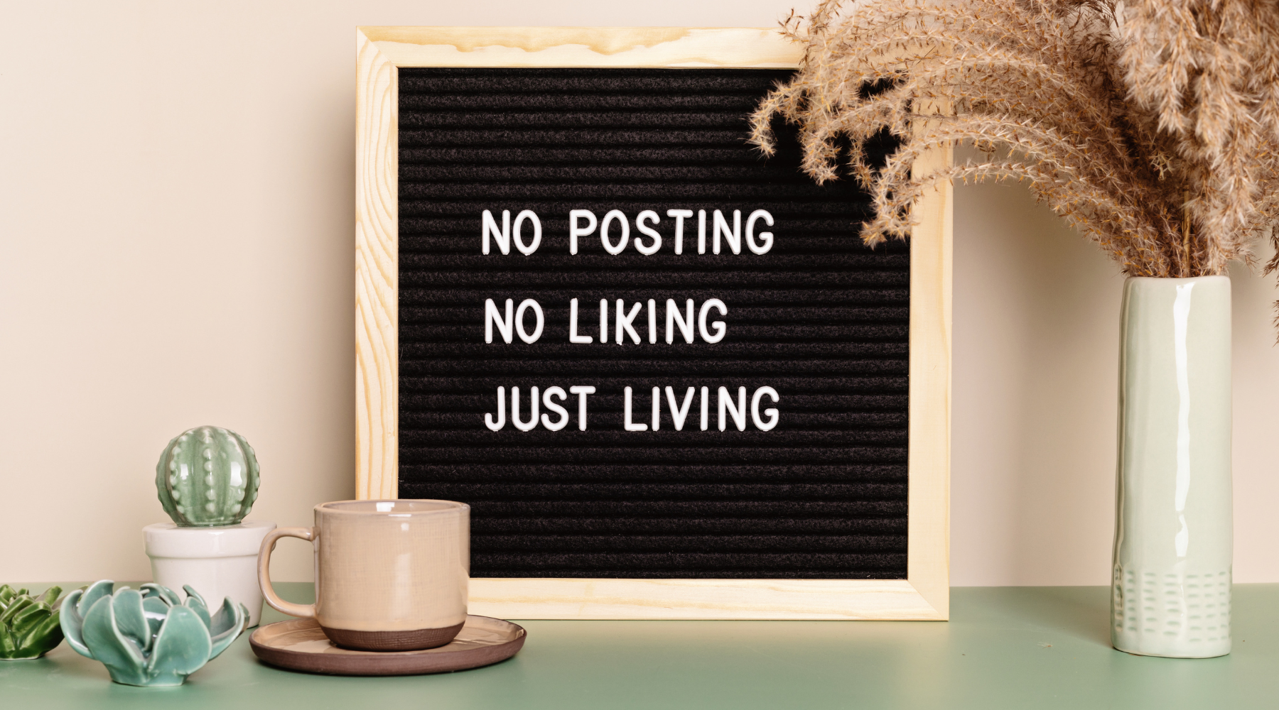 Black felt letter board with wooden frame displaying the text: NO POSTING NO LIKING JUST LIVING, surrounded by a ceramic cactus, a cup on a saucer, and a tall vase with dried flowers.