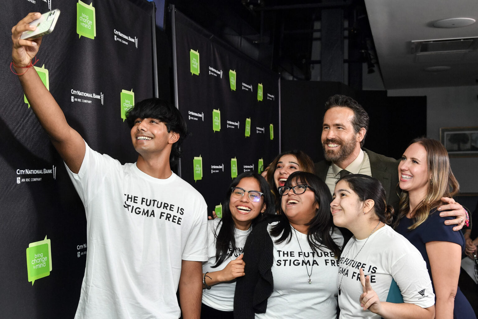 A group of six smiling people, including young adults wearing white shirts with the text 'THE FUTURE IS STIGMA FREE', taking a selfie in front of a backdrop with repeated logos 'bring change to mind' and 'City National Bank AN RBC COMPANY'.