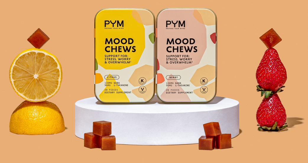 Two tins of PYM Mood Chews dietary supplements labeled Citrus and Berry flavors, each with 130mg GABA and 90mg L-Theanine, displayed on a white pedestal with citrus fruit and strawberry stacks and chew cubes around them.