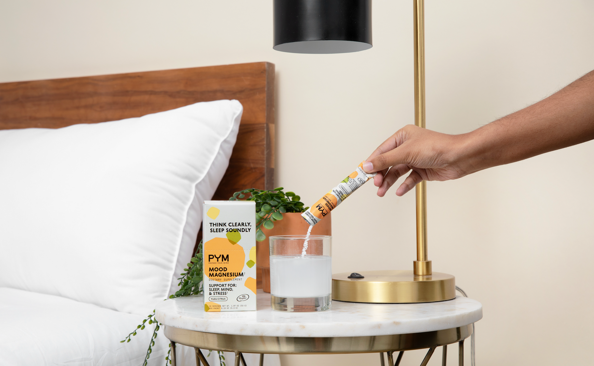Hand pouring PYM Mood Magnesium powder into a glass of water on a bedside table with a lamp and plant; product box text: THINK CLEARLY, SLEEP SOUNDLY, PYM MOOD MAGNESIUM, DIETARY SUPPLEMENT, SUPPORT FOR: SLEEP, MIND, & STRESS, YUZU CITRUS.