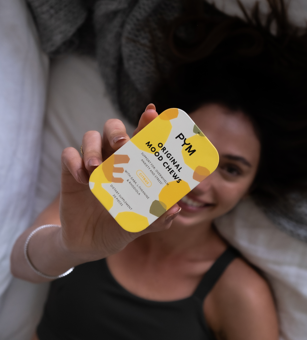 Woman holding a yellow tin labeled 'PYM ORIGINAL MOOD CHEWS SUPPORT FOR OVERWHELM ANXIETY AND STRESS CITRUS WITH GABA L-THEANINE & RHODIOLA DIETARY SUPPLEMENT 20 PIECES'