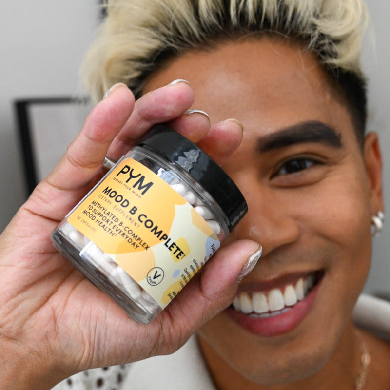 Person holding a jar of PYM Mood B Complete dietary supplement while smiling with label text: 'PYM MOOD B COMPLETE DIETARY SUPPLEMENT METHYL B COMPLEX TO SUPPORT EVERYDAY MOOD HEALTHY'