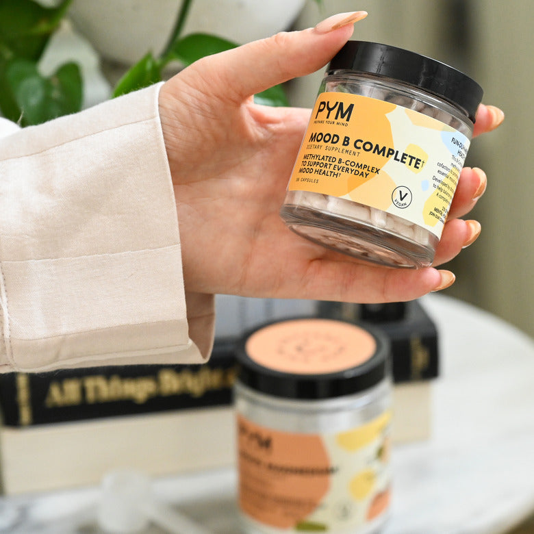 Hand holding a jar of PYM Mood B Complete dietary supplement capsules with label text: 'PYM, Mood B Complete, Dietary Supplement, Methylated B-Complex to Support Everyday Mood Health, Vegan, 30 Capsules'