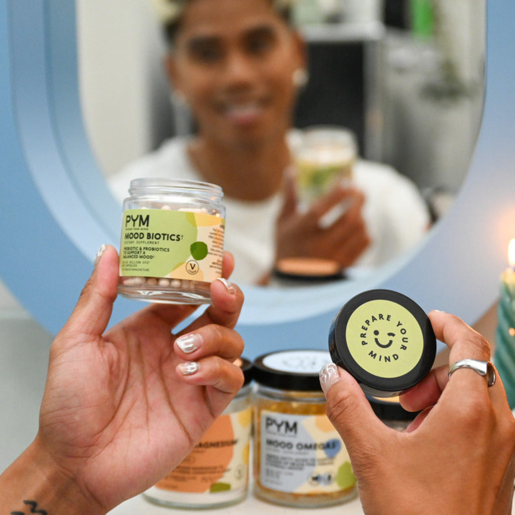 Person holding a jar of PYM Mood Biotics dietary supplement and the lid with text 'Prepare Your Mind'. Two other PYM supplement jars are visible on the table.