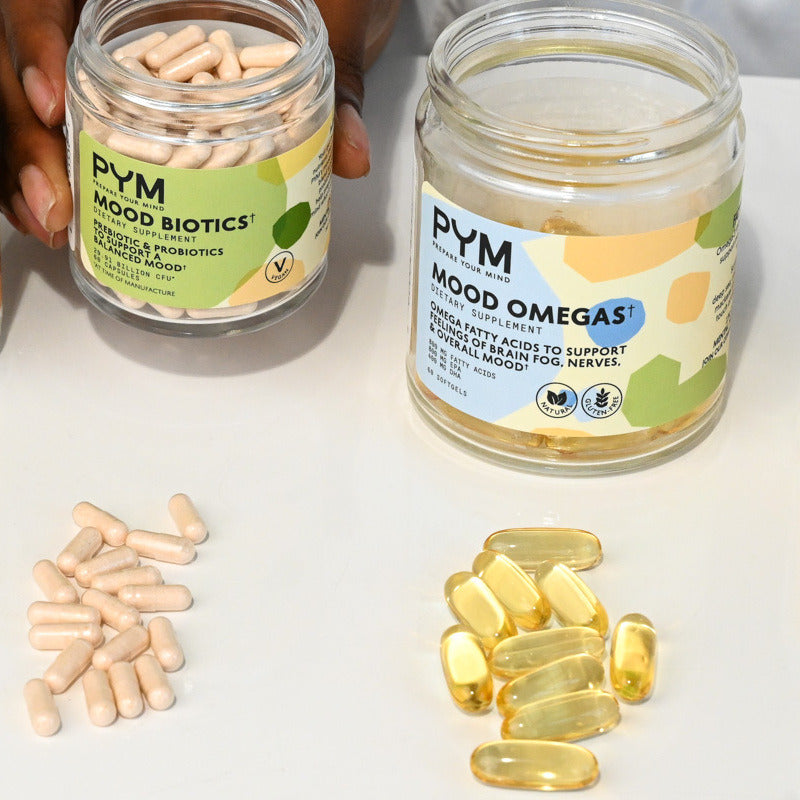 Two jars of dietary supplements labeled 'PYM MOOD BIOTICS†' and 'PYM MOOD OMEGAS†' with beige capsules and yellow softgel capsules spilled in front of them.