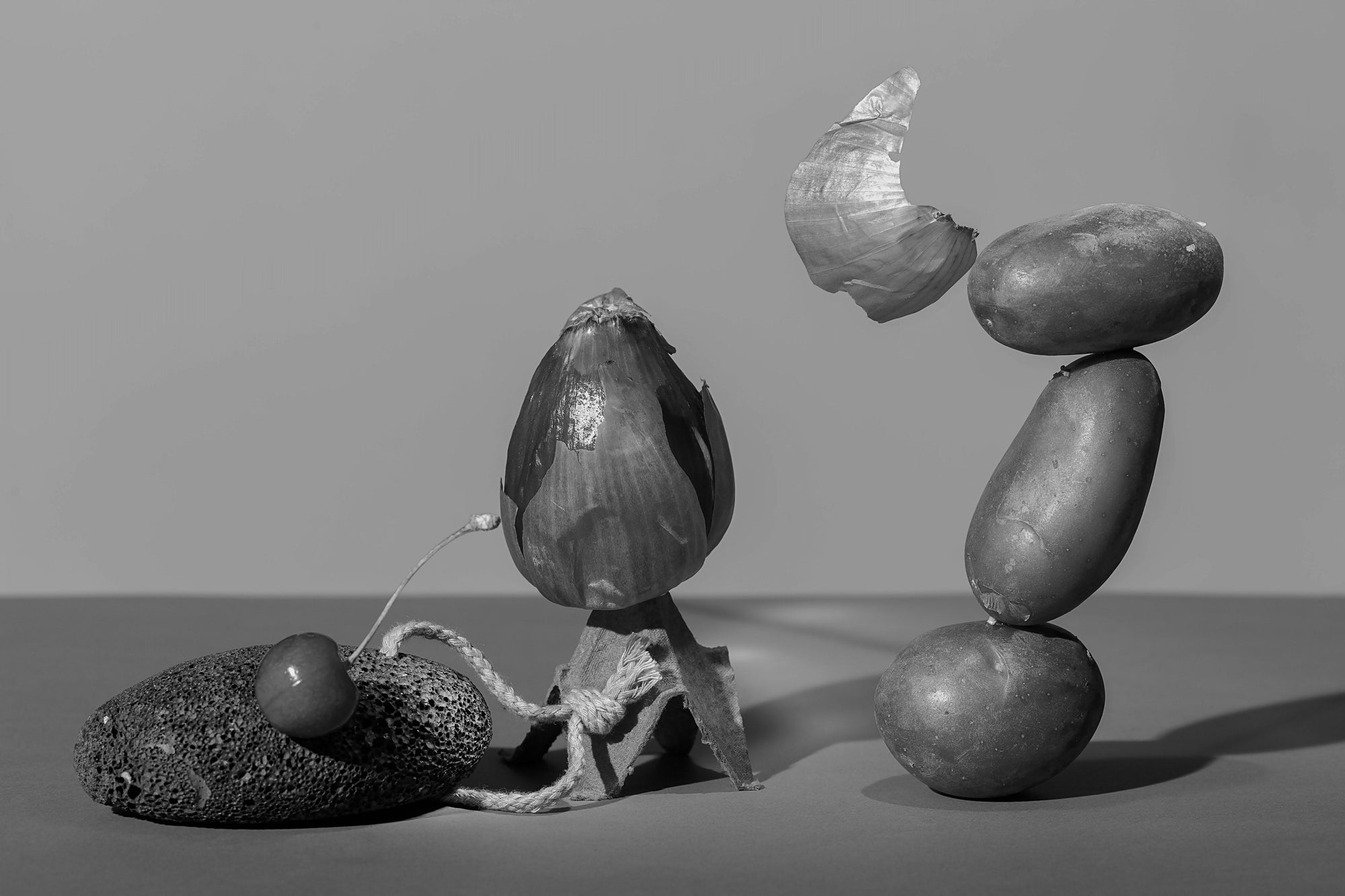 Still life composition with a cherry on a porous rock connected by a string to an eggplant with its peel partially removed, and three potatoes stacked vertically with a piece of onion peel balanced on top.