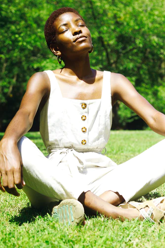 A person sitting cross-legged on grass outdoors with eyes closed, appearing relaxed and peaceful, wearing a white sleeveless outfit with buttons.