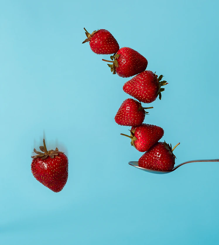 Six red strawberries, five stacked on a spoon and one falling against a light blue background.