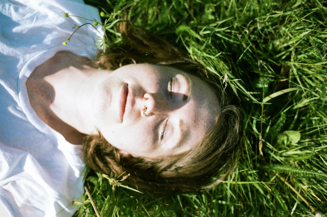 Person with closed eyes lying on green grass, wearing a white shirt, with sunlight casting shadows on their face.