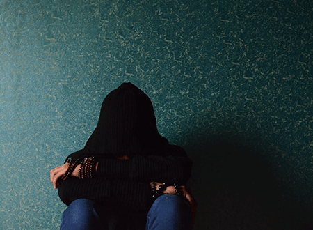 Person wearing a black hoodie sitting with head down against a textured blue wall, conveying sadness or depression