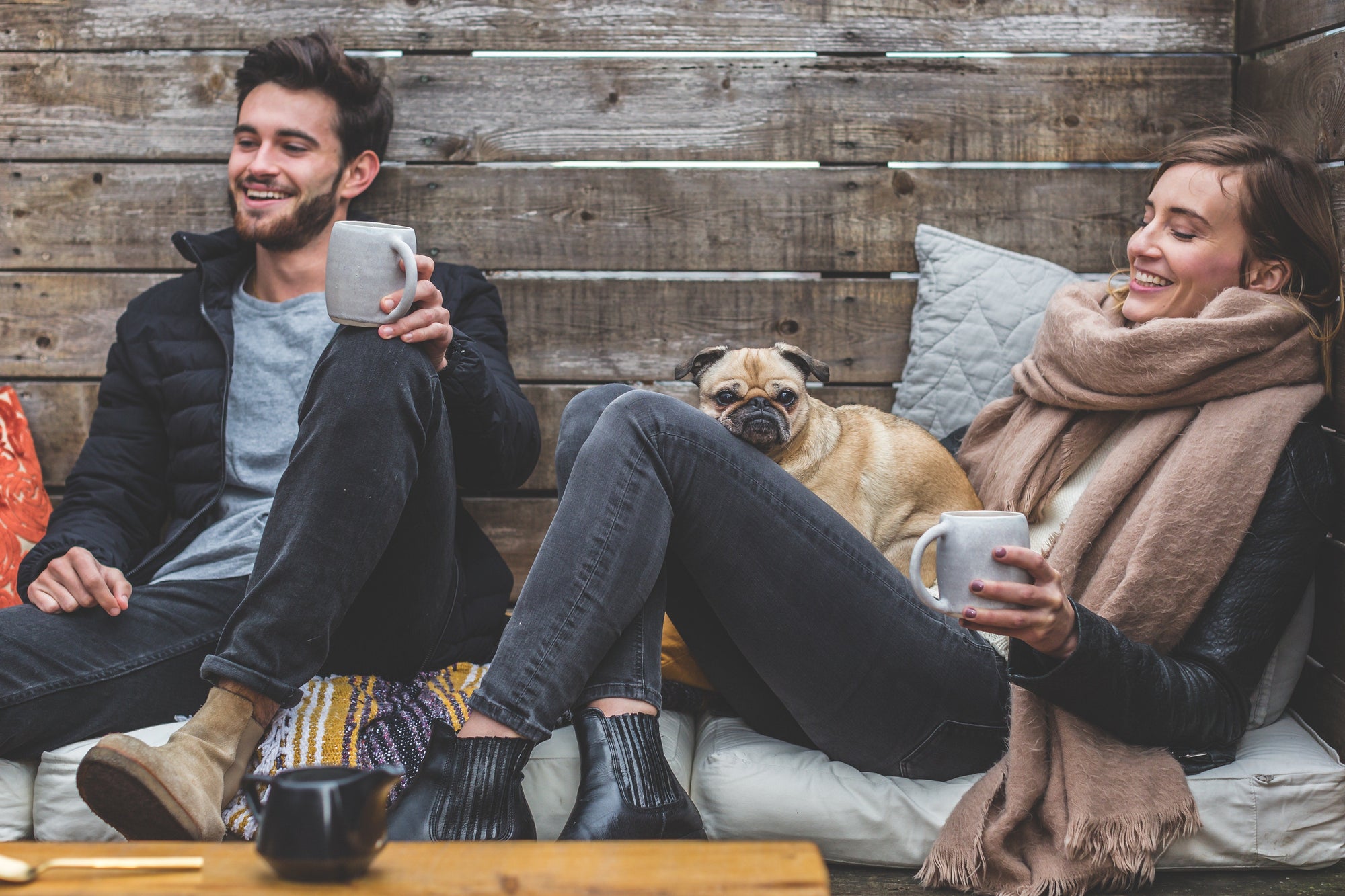 Two people sitting on a cushioned bench against a wooden wall, smiling and holding mugs, with a pug resting on the woman's lap. Text in image: '5 Ways To Make Someone Happy Today'.