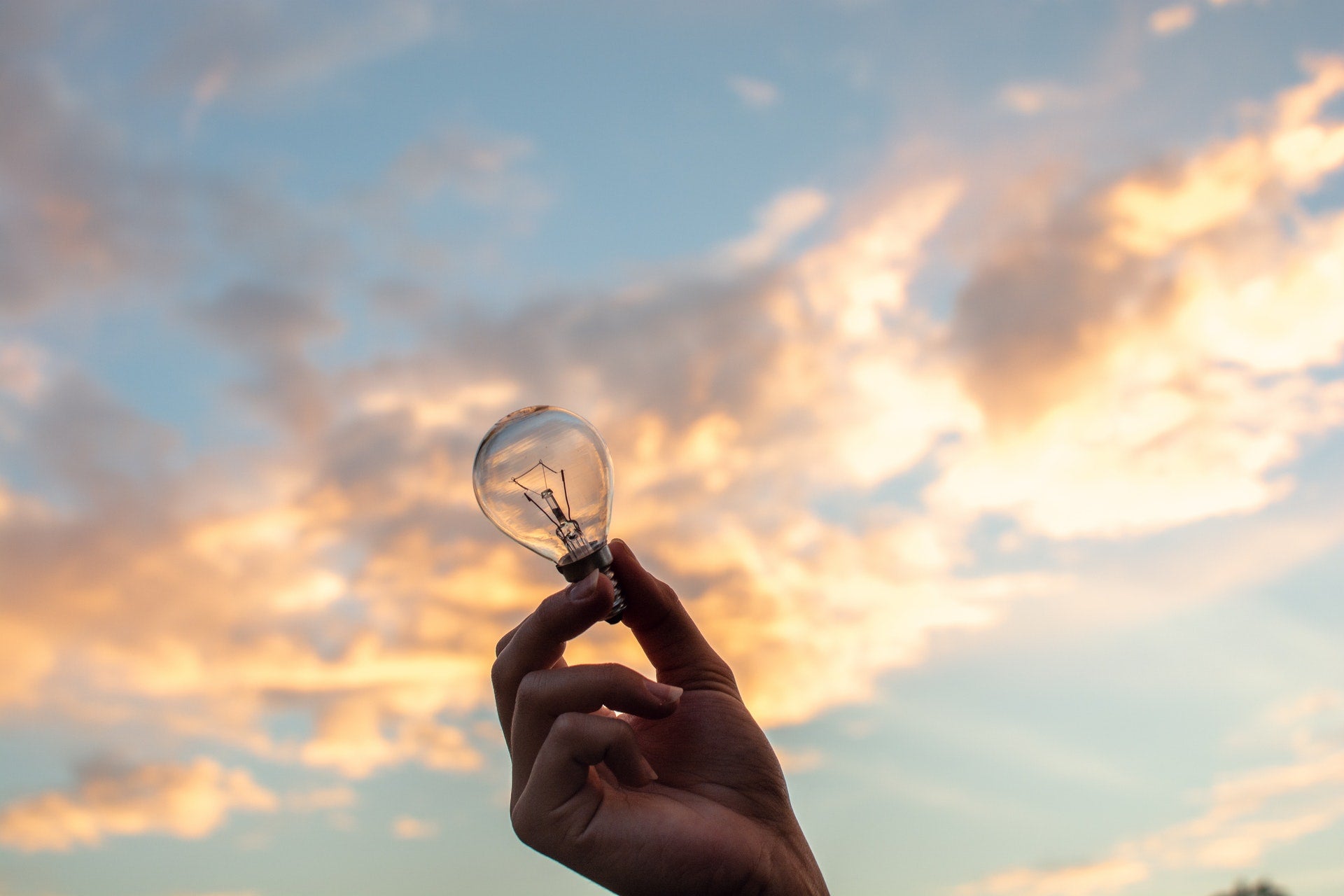 Hand holding a clear light bulb against a colorful sunset sky with clouds