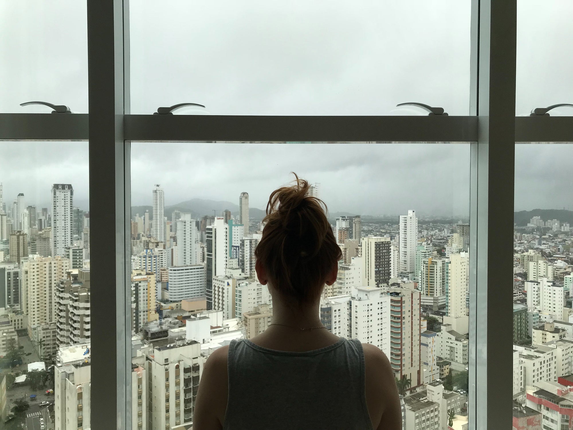Person with hair in a bun looking out a large window at a city skyline under cloudy sky