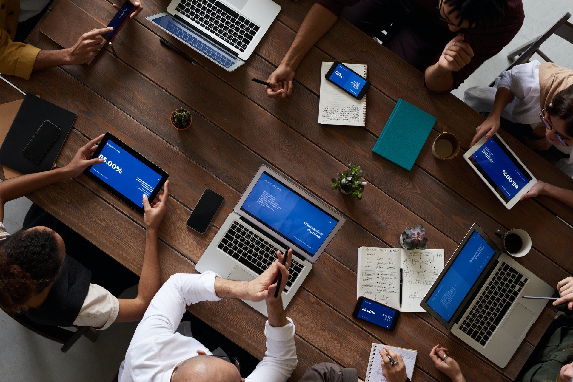 Overhead view of a group meeting around a wooden table with laptops, tablets, and phones showing blue presentation slides. Visible text includes 'Discussion Outline', '85.00%', and handwritten notes on notebooks.