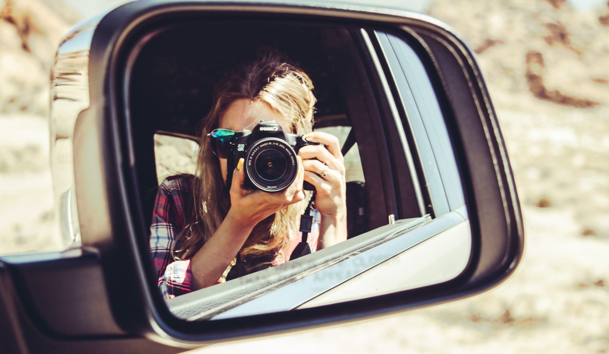 Person taking a photo with a camera reflected in a car's side mirror in a desert setting