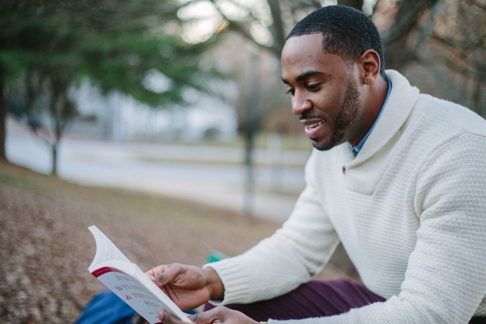 Man in a white sweater reading a book outdoors; visible text on book cover includes 'WIN PEACE & INFLUENCE PEOPLE'