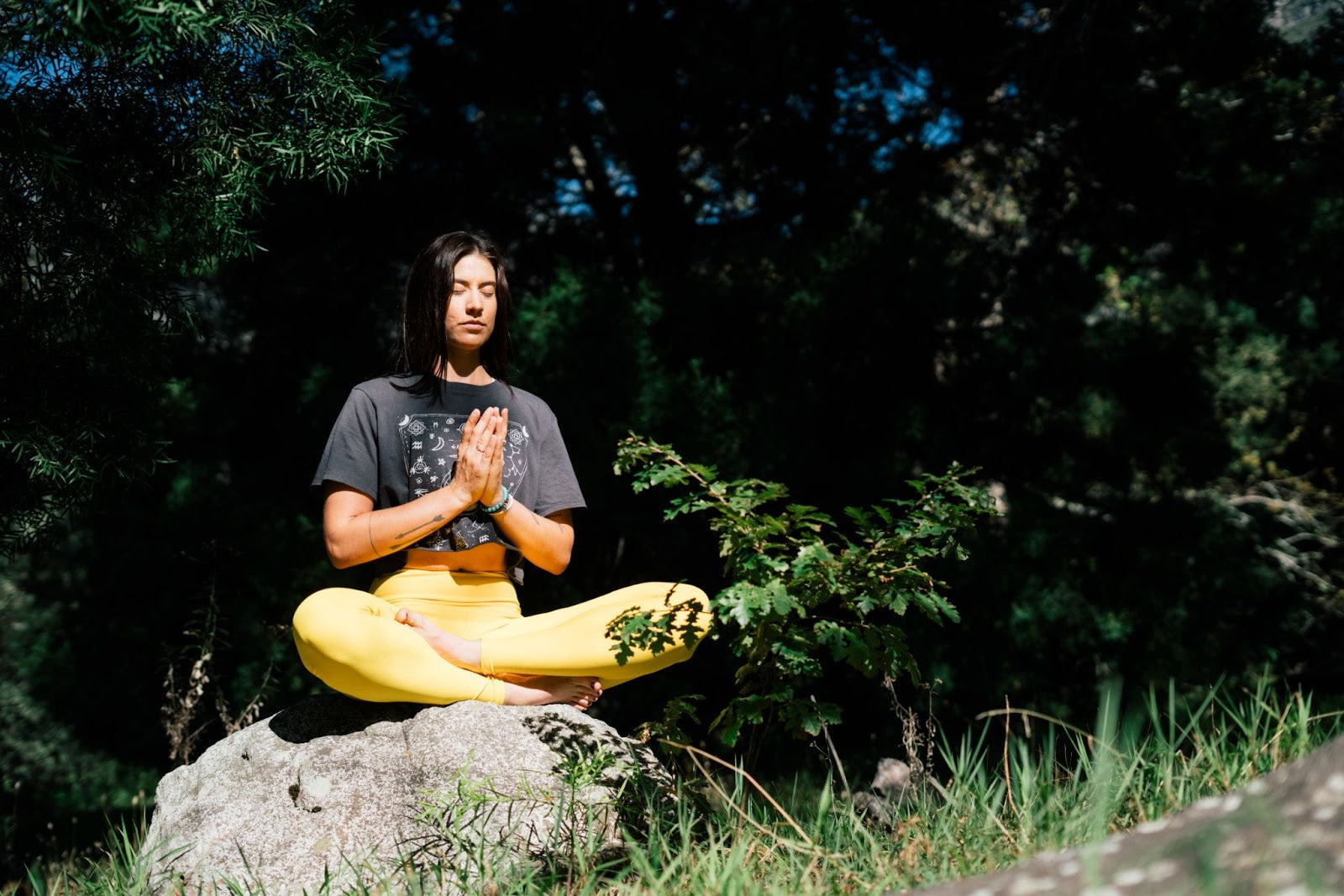 Woman meditating outdoors on a rock with eyes closed, wearing yellow leggings and a dark t-shirt, surrounded by greenery.