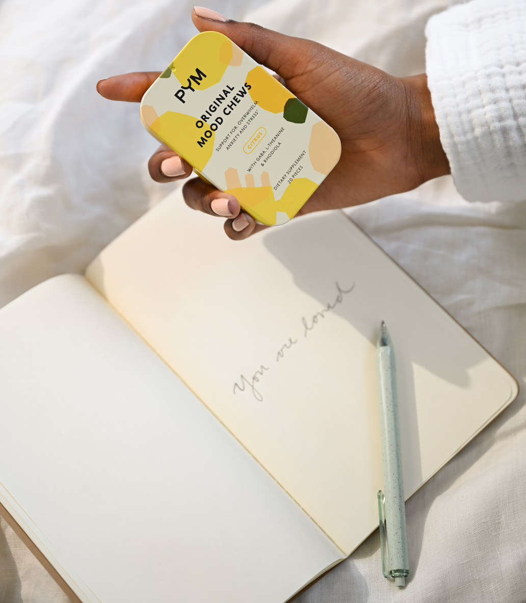 Hand holding a tin of PYM Original Mood Chews over an open notebook with handwritten text 'You are loved' and a pen lying on it.
