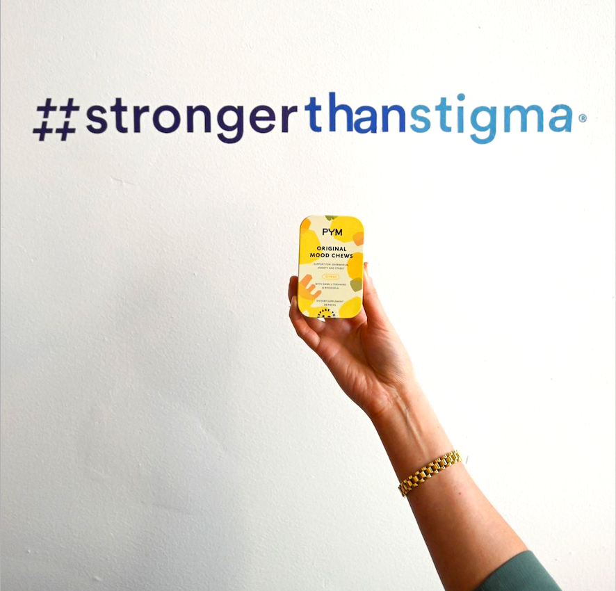 Hand holding a yellow tin labeled 'PYM Original Mood Chews' under the text '#strongerthan stigma' on a white wall.
