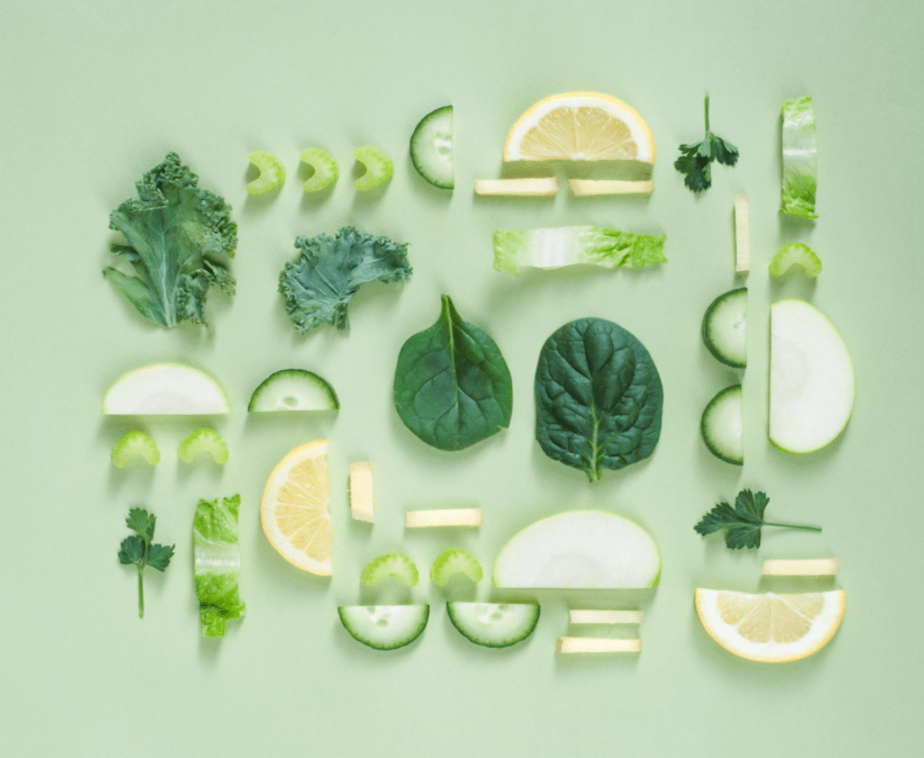 Various green leafy vegetables and sliced fruits including kale, spinach, cucumber, lemon, celery, and parsley arranged neatly on a green background.