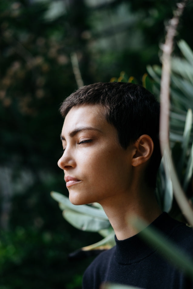 Profile of a person with short dark hair and eyes closed, surrounded by green foliage.