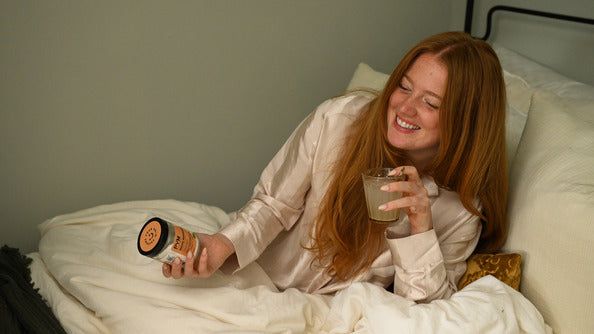 Smiling woman in bed holding a jar and a glass, suggesting relaxation and mental hygiene