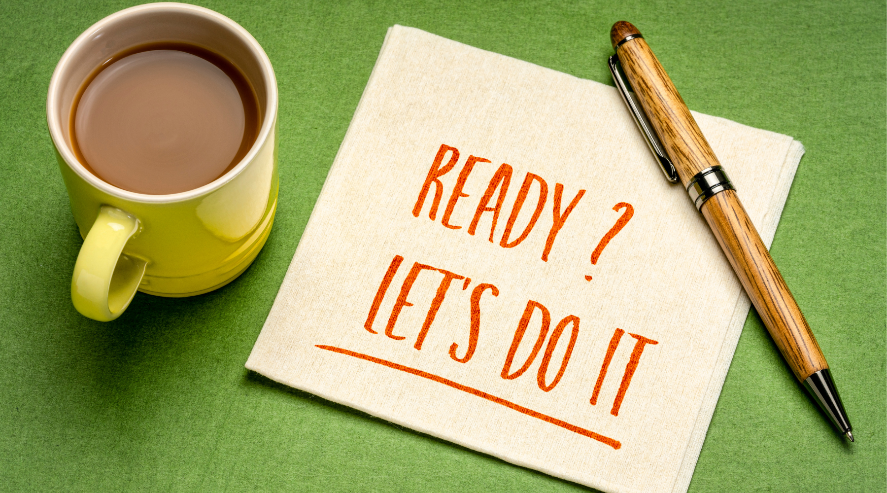Yellow coffee cup, wooden pen, and napkin with text 'READY? LET'S DO IT' on green surface